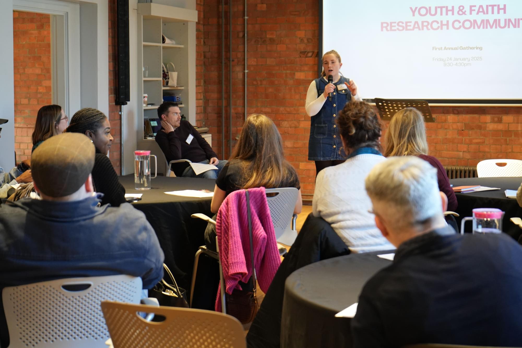 A woman speaks at a podium in front of a room of seated people. The slide behind her reads "Youth & Faith Research Community" with a date in January 2023. The attendees sit at round tables with black tablecloths in a brick-walled room.