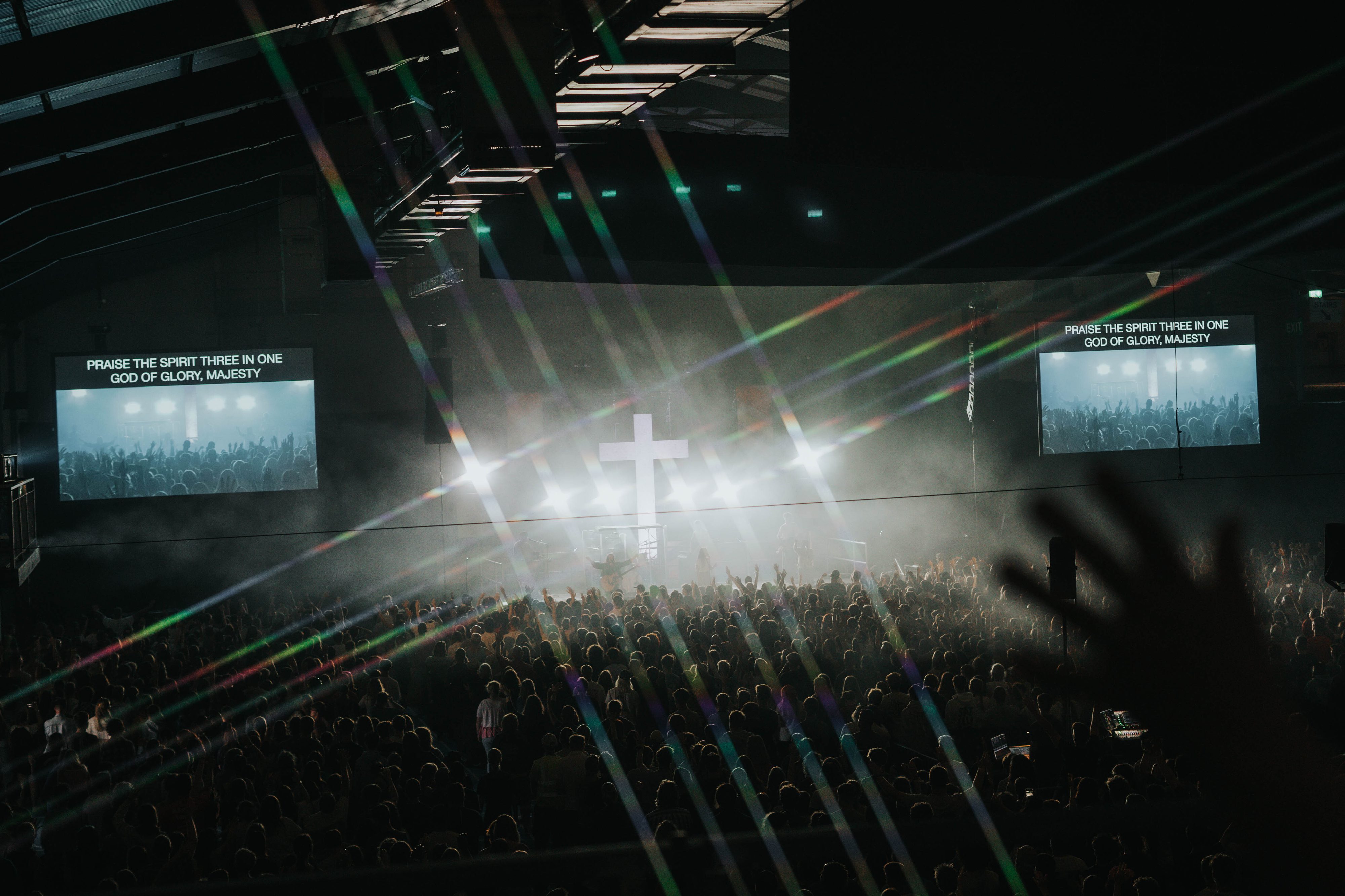 A large crowd gathers at a concert or worship event in a dark indoor venue. Bright stage lights and a white cross are visible on stage, with two screens displaying song lyrics above the audience.